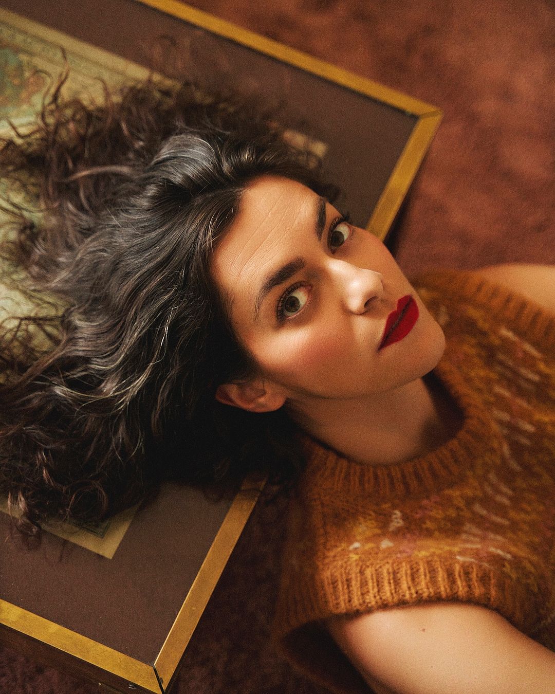 Eye-level close-up shot of an adult woman with dark curly hair, red lipstick and a knit top lying on a picture frame and dark brown surface.