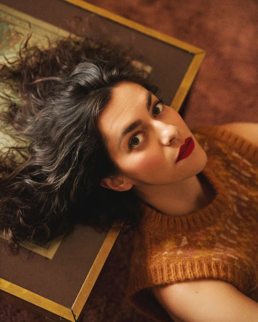 Eye-level close-up shot of an adult woman with dark curly hair, red lipstick and a knit top lying on a picture frame and dark brown surface.