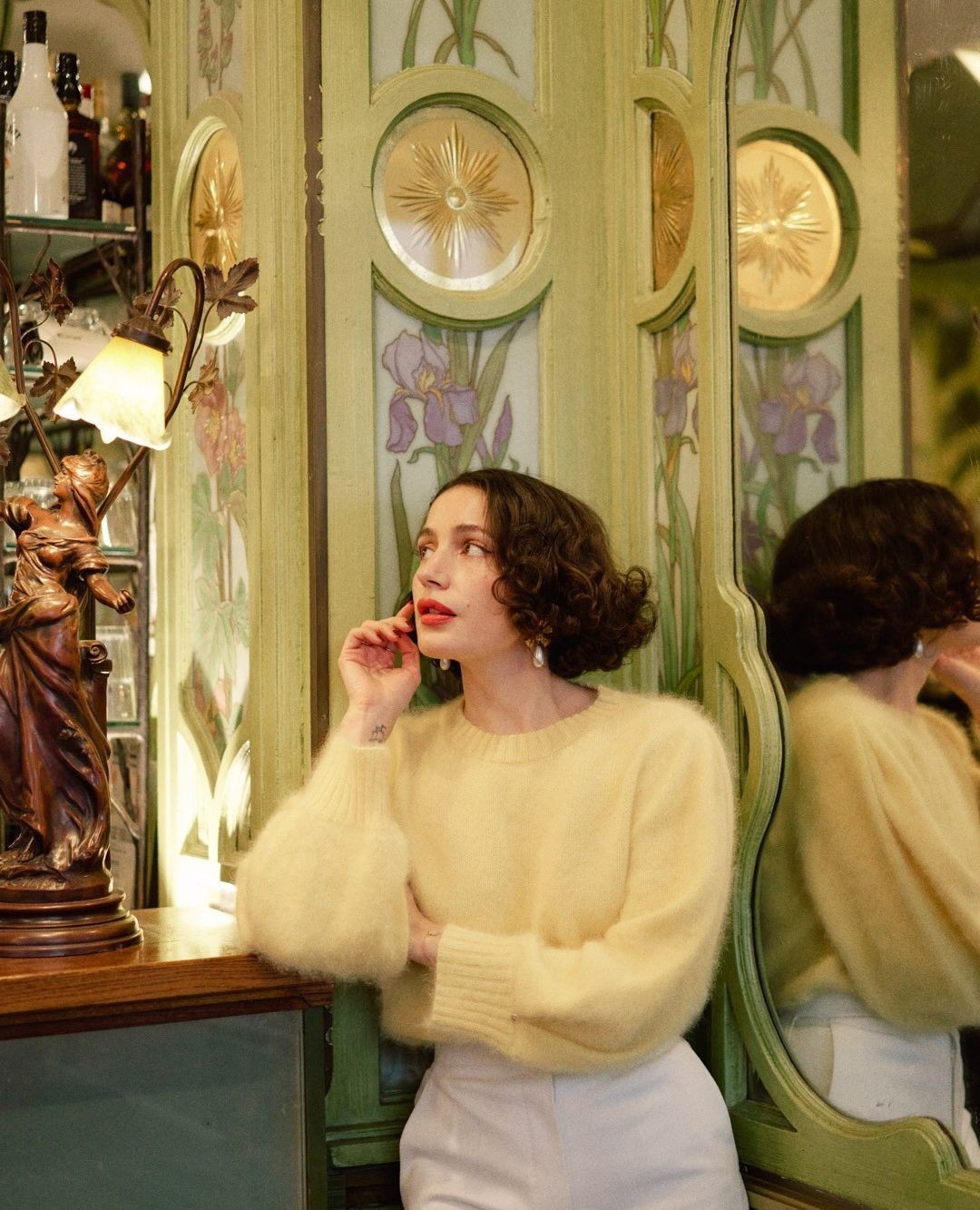 Brunette woman with curly hair poses in a yellow sweater and white pants in front of a floral-painted green wall and mirror.