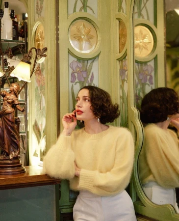 Brunette woman with curly hair poses in a yellow sweater and white pants in front of a floral-painted green wall and mirror.