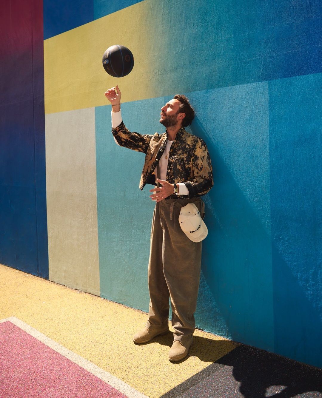 Adult male stands against a vibrant geometric wall, balancing a basketball on his finger in bright sunlight. Casual fashion attire.