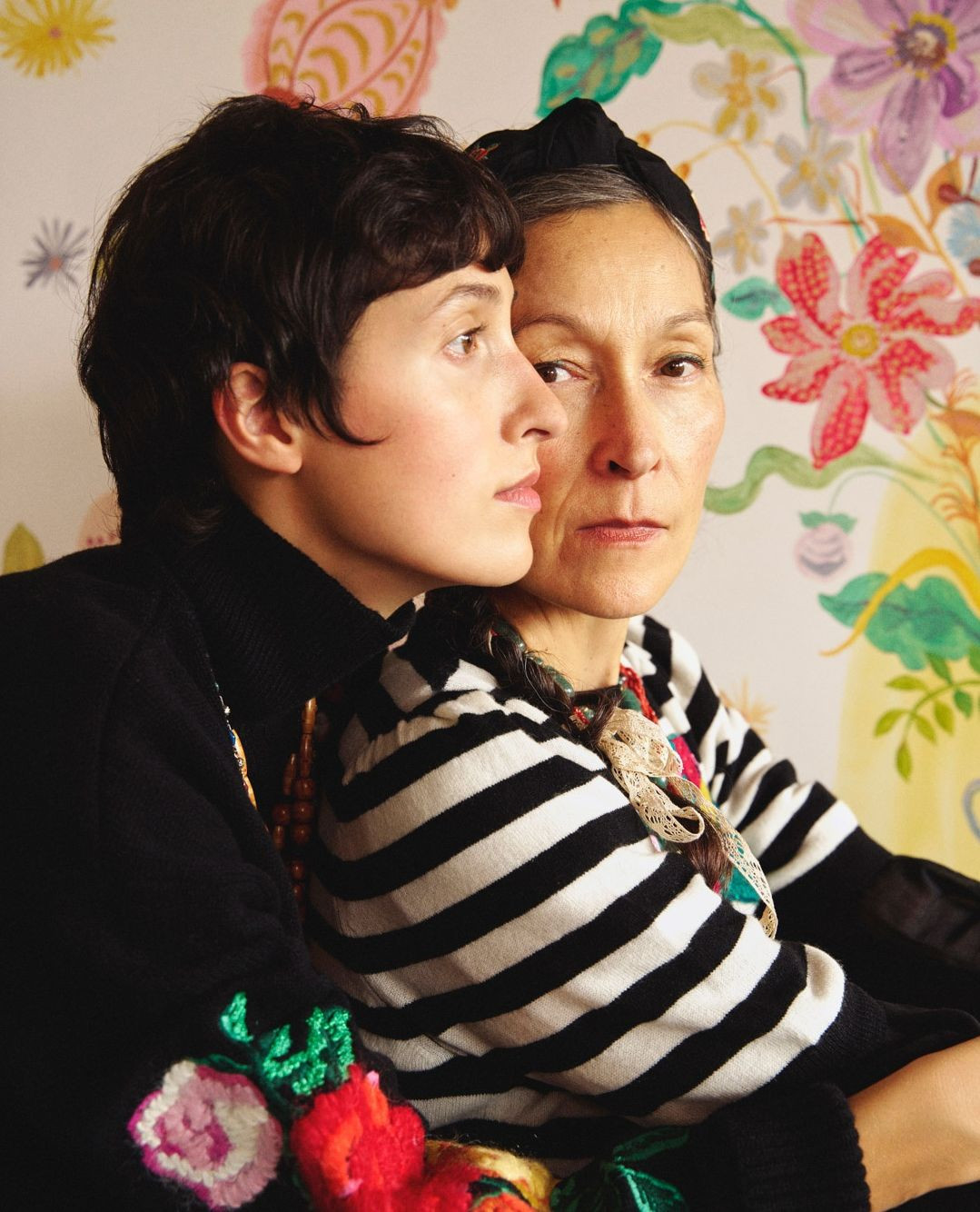 Two women, one younger and one older, pose indoors with a floral wallpaper backdrop, wearing black and striped tops, looking serene.