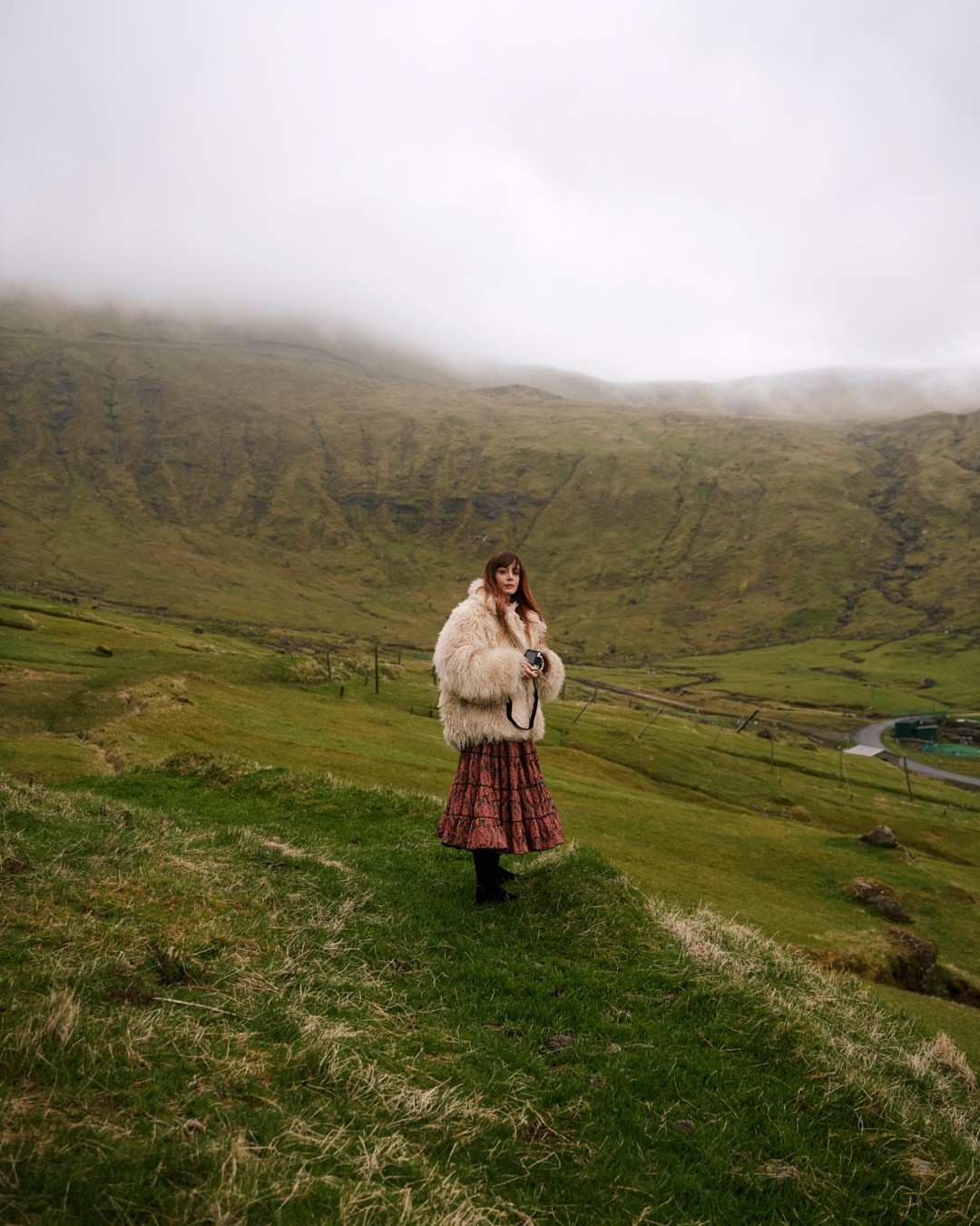 A young woman in a furry coat and patterned skirt stands in a grassy field, holding a camera, with a misty mountain backdrop.