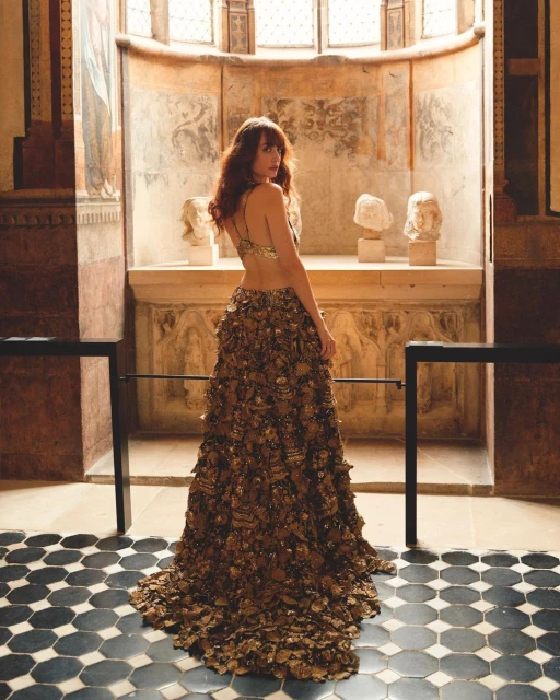 Woman in an elaborate gold gown standing in a historic hall with marbled floors, antique busts, frescoes, and natural light from arched windows.