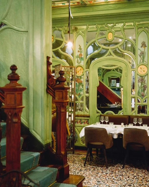 Interior view of an elegant restaurant features green walls, wooden staircase, tables set with glasses and plates, and a patterned floor.