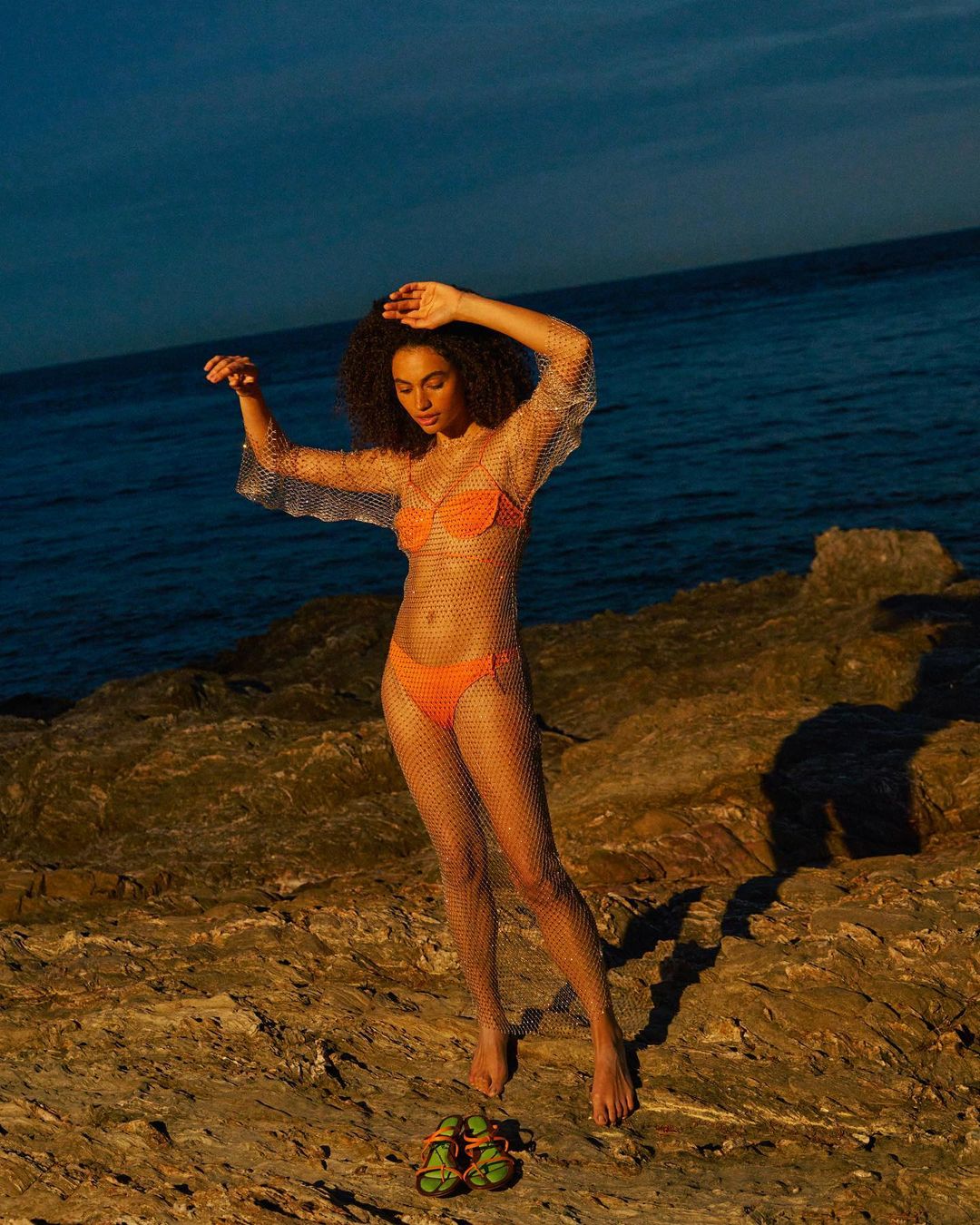 Woman in orange swimwear and a mesh dress poses on rocks near the ocean in the evening light with sandals on the ground.