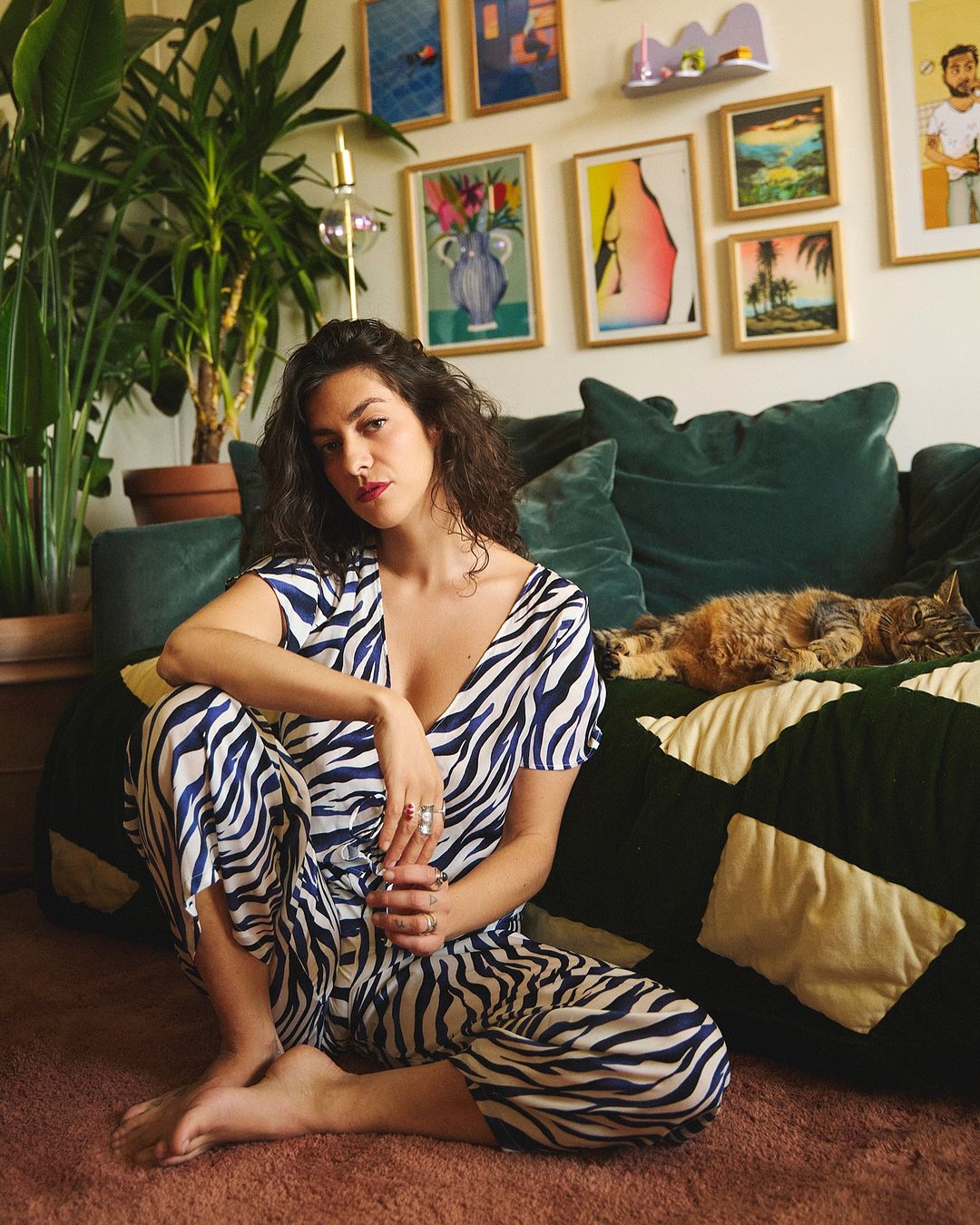 A woman in a zebra-striped jumpsuit sits barefoot on a brown carpet with a cat on a dark green couch, surrounded by colorful wall art.