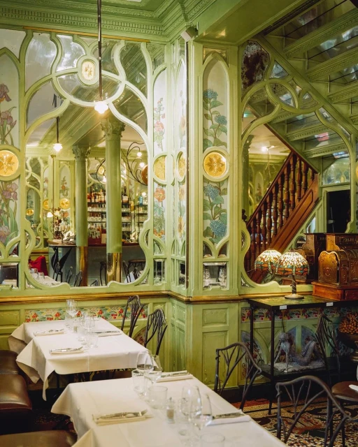 Interior of an ornate restaurant with art nouveau details, dining tables, chairs, and a bar area with glassware and bottles.