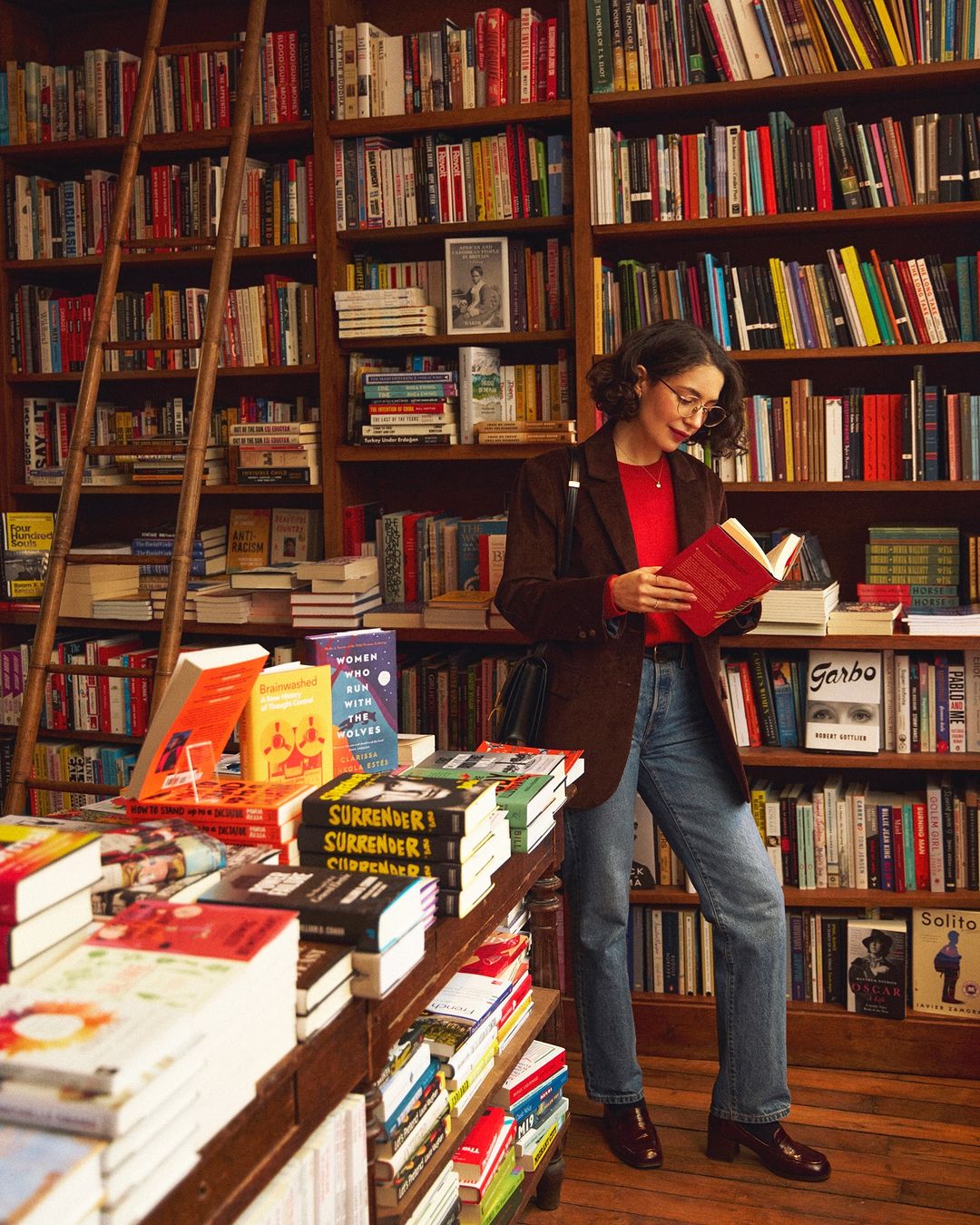Adult female reads a book in a bookstore filled with tall bookshelves, wearing a red shirt, brown blazer, and jeans in a full shot.