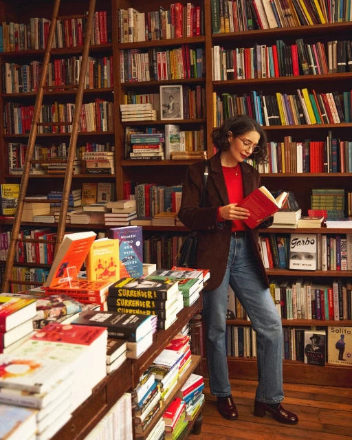 Adult female reads a book in a bookstore filled with tall bookshelves, wearing a red shirt, brown blazer, and jeans in a full shot.