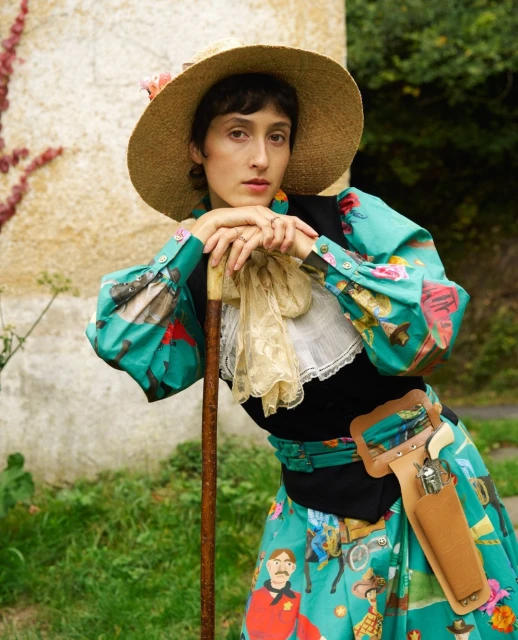 Woman in vintage dress, sun hat, leans on wooden cane outside. She has dark hair, fair skin, and is wearing a black vest over the gown.