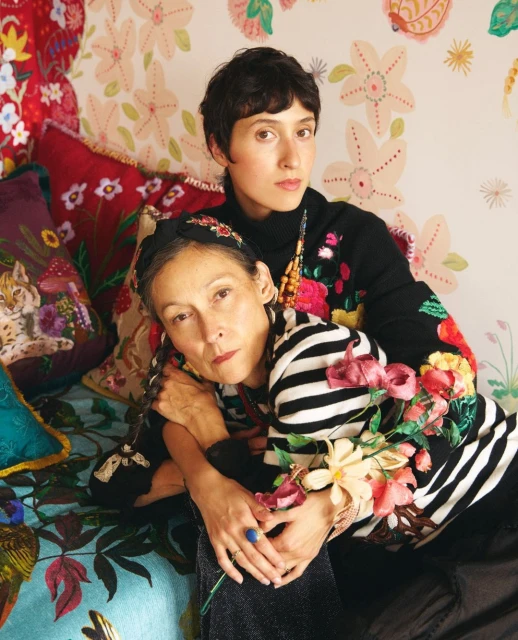 Two women embrace indoors on a floral bed with colorful floral wallpaper. One woman wears a striped shirt, the other a black turtleneck with floral accents.