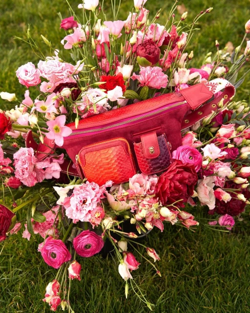 Red fanny pack surrounded by pink, white, and red flowers laying on green grass in an eye-level outdoor shot showcasing fashion.
