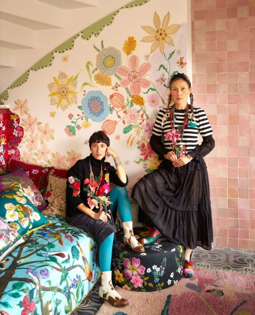 Two stylish women pose indoors against a colorful floral backdrop, seated on a patterned couch and ottoman in a vibrant, artistic setting.