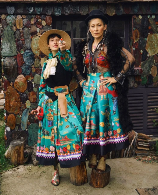 Two women in brightly patterned vintage dresses pose outdoors in front of a stone wall, one holding a toy gun, striking a playful pose.