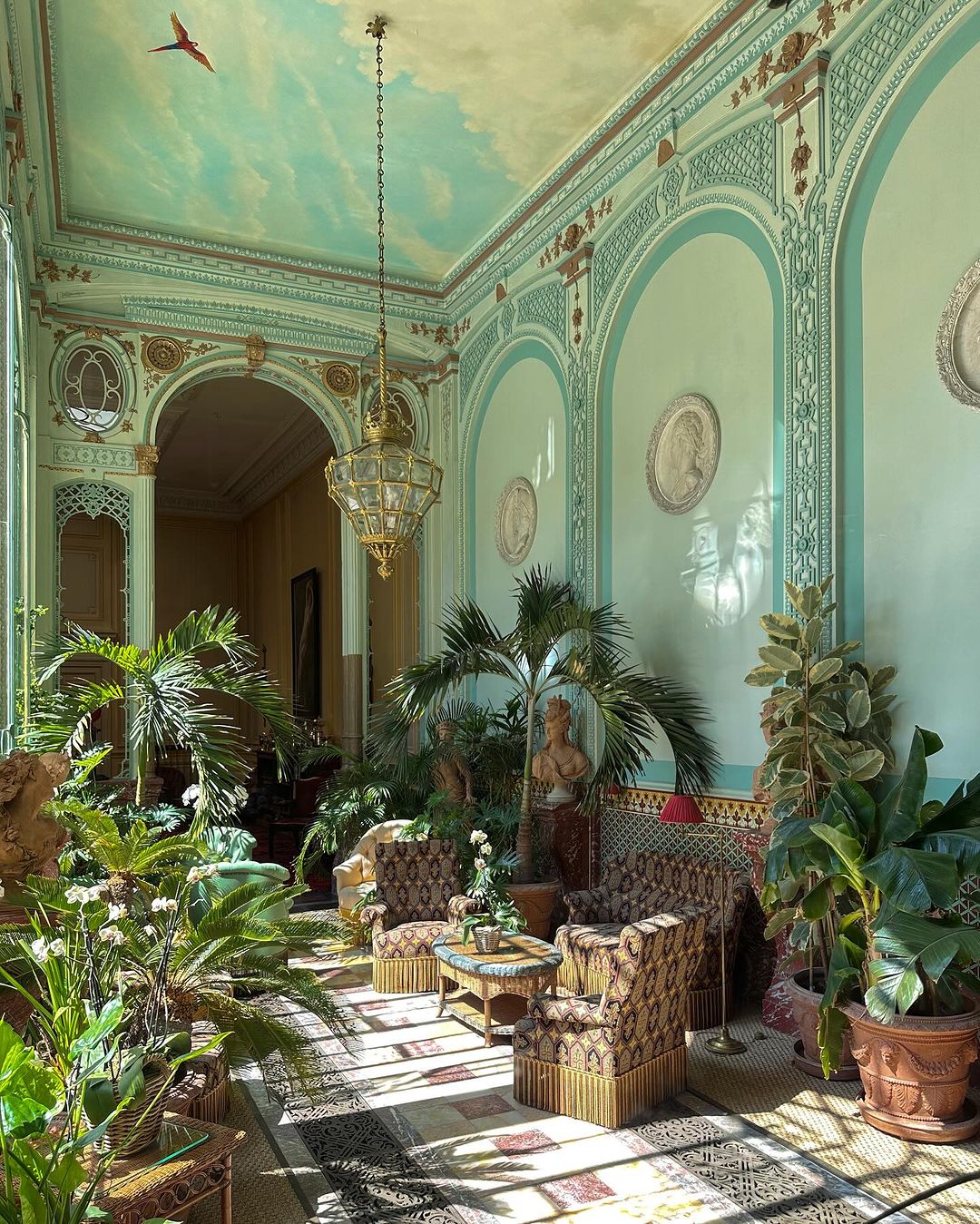 Opulent conservatory with patterned chairs, abundant potted plants, decorative chandelier, and arched architectural details under a painted sky ceiling.