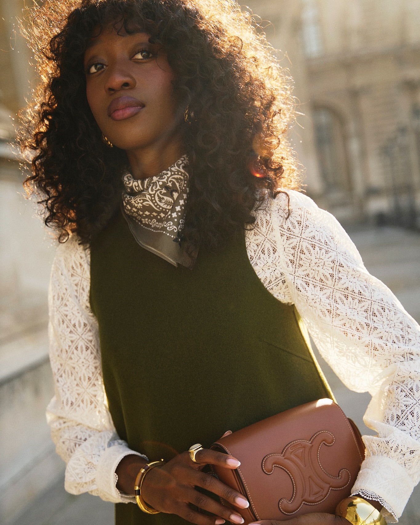 Stylish Black woman poses in an olive dress with white lace sleeves, accessorized with gold jewelry and a brown leather handbag against building.