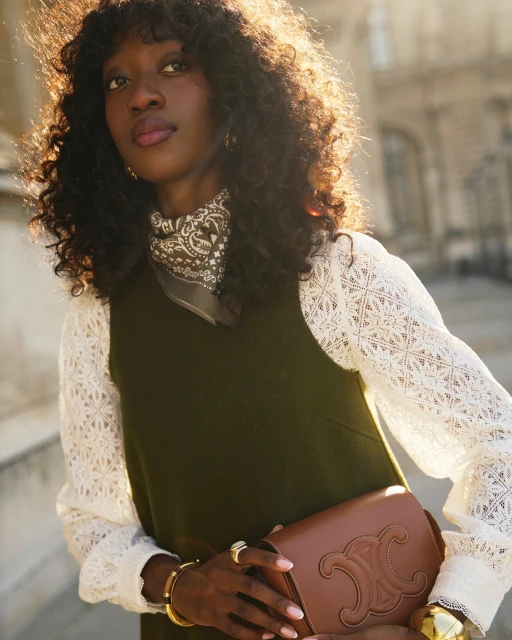 Stylish Black woman poses in an olive dress with white lace sleeves, accessorized with gold jewelry and a brown leather handbag against building.