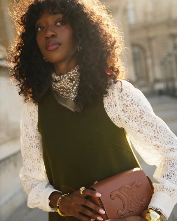 Stylish Black woman poses in an olive dress with white lace sleeves, accessorized with gold jewelry and a brown leather handbag against building.