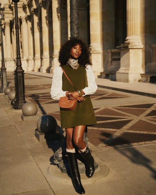 A stylish Black woman poses in an olive green dress and black boots in an urban setting with classical architecture in the background.