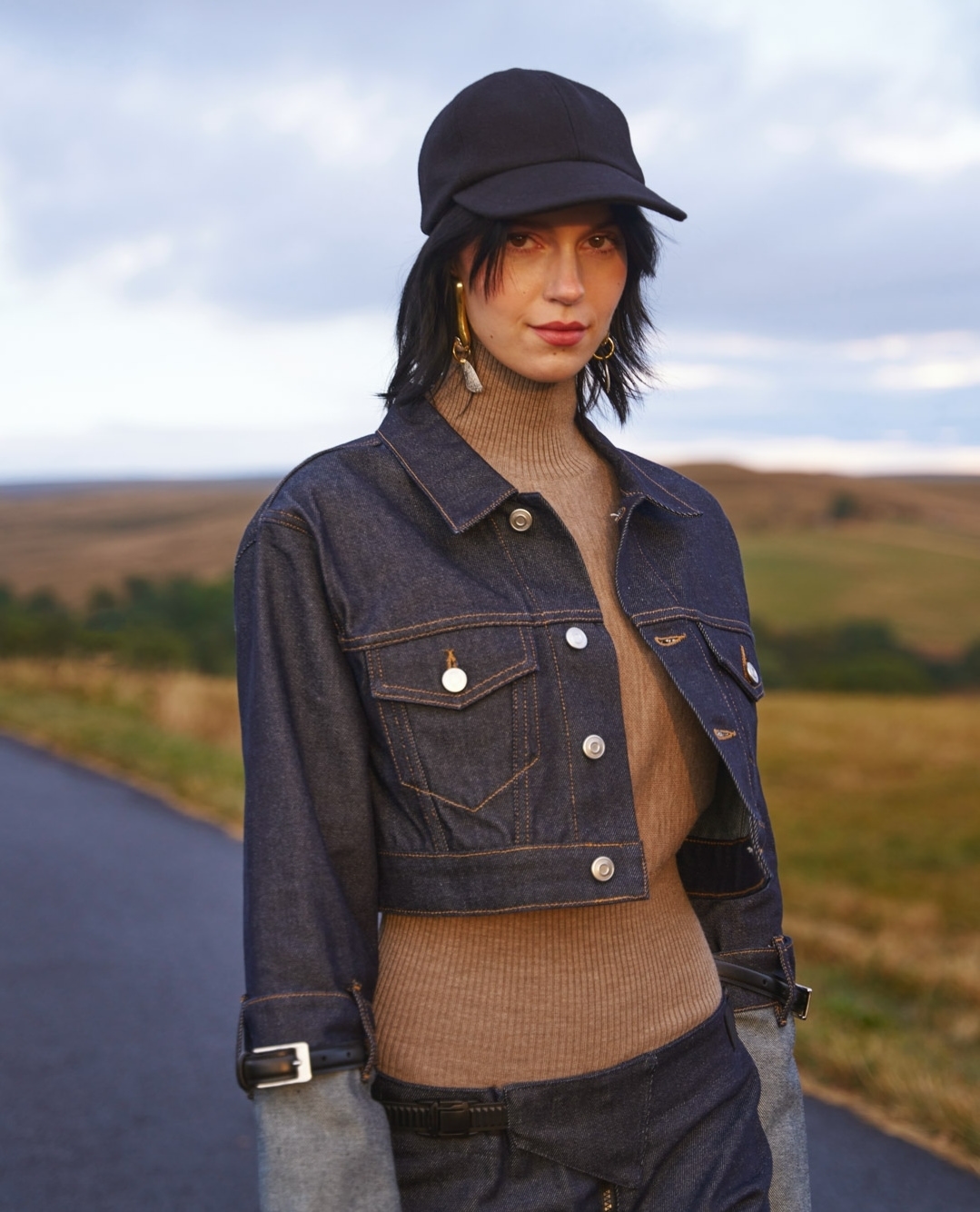 A woman with dark hair, wearing a cap and denim jacket, poses on a rural road with grassy hills and cloudy skies in the background.