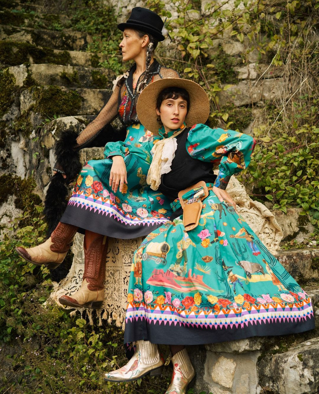 Two women in Western-style fashion sit on stone steps surrounded by greenery. They wear hats, vests, long floral skirts, and cowboy boots.