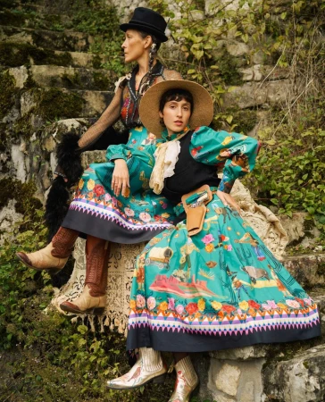 Two women in Western-style fashion sit on stone steps surrounded by greenery. They wear hats, vests, long floral skirts, and cowboy boots.