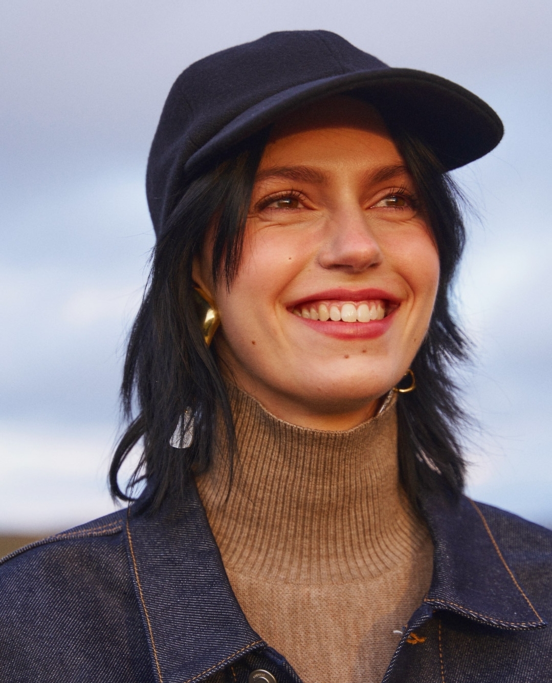 Smiling young woman with dark hair in a cap and denim jacket, set against a blurred background of clouds and earth tones.