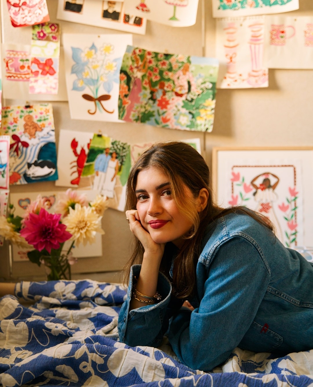 Young woman in denim jacket leans on hand while surrounded by floral artwork in an indoor setting with a patterned blue and white blanket.