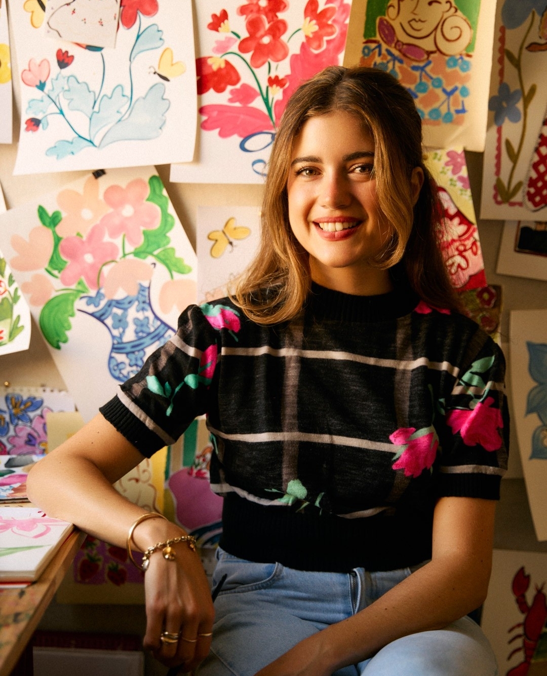 Smiling woman in floral top sits in front of floral art on a wall. A cheerful and creative indoor portrait.
