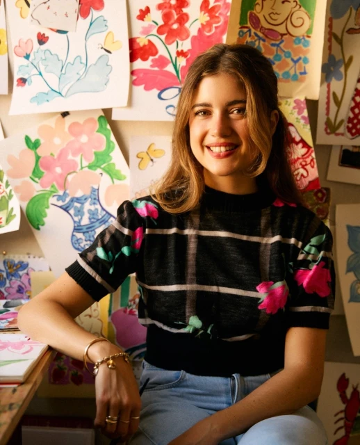 Smiling woman in floral top sits in front of floral art on a wall. A cheerful and creative indoor portrait.