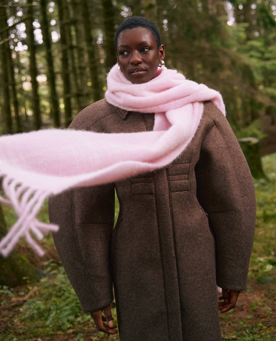 A dark-skinned woman in a brown coat and a pink scarf stands in a forest, looking off to the side in a medium shot.