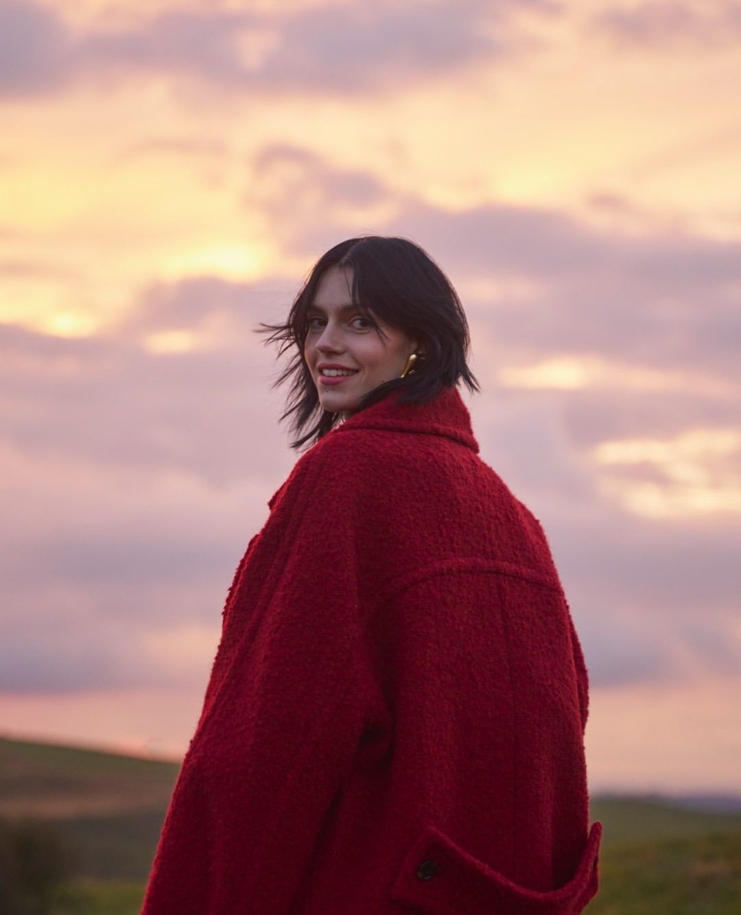 Young woman in a red coat stands in a field at dusk, looking back over her shoulder with a smile, against a cloudy sky during sunset.