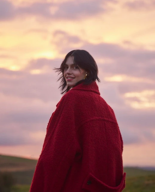 Young woman in a red coat stands in a field at dusk, looking back over her shoulder with a smile, against a cloudy sky during sunset.