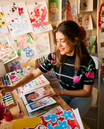Female artist smiles in her studio, surrounded by colorful watercolor paintings, wearing a patterned sweater and a flower in her hair.