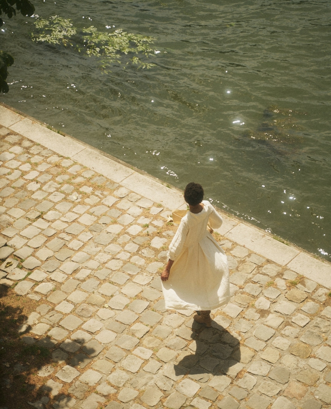 Woman in a white dress stands at the edge of a cobblestone walkway near a body of water on a sunny day with tree shadow.
