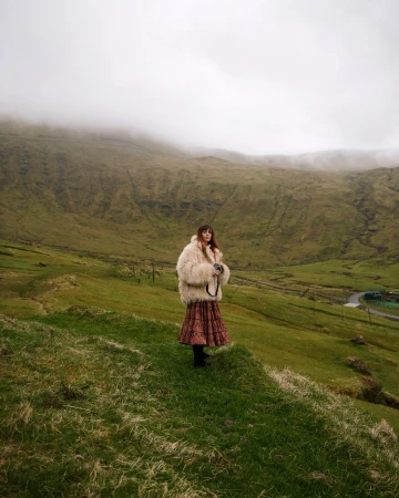 Woman in a fluffy cream coat and patterned skirt holding a camera in a misty green valley