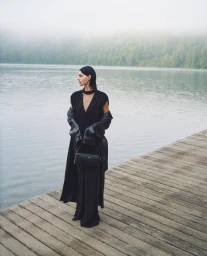 Model in black draped dress and leather coat standing on a wooden dock by a misty lake, photographed by Pauline Darley.