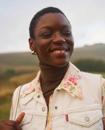 Portrait of a smiling black woman wearing a white denim vest over a floral shirt and brown turtleneck against an outdoor landscape.