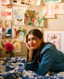 Young woman in denim jacket leaning on a patterned blue blanket with colourful floral illustrations pinned on the wall behind her.