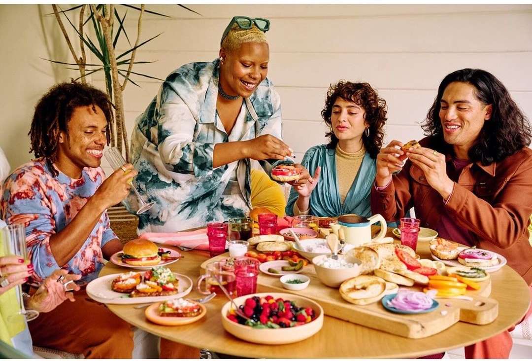 Diverse group of young adults enjoying a brunch-style meal at a round table, with a variety of food and drinks in a bright, indoor setting.