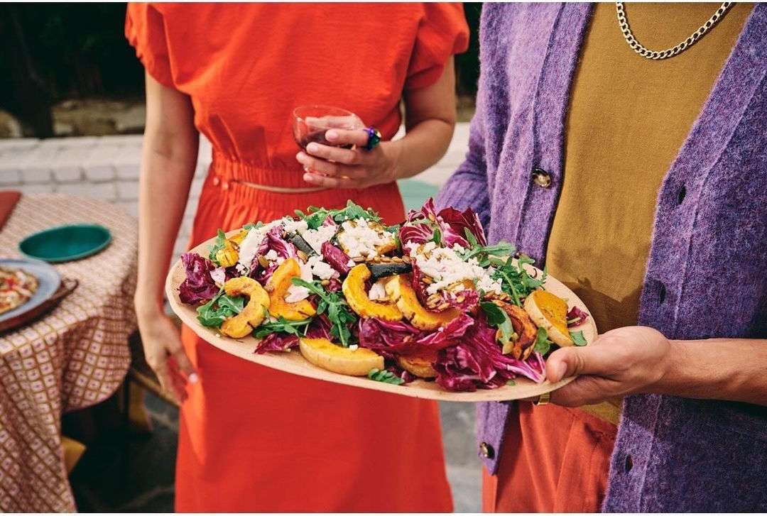 A person holds a platter of butternut squash salad with arugula and cheese, while another person holds a wine glass in an outdoor setting.