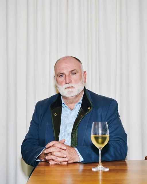 An adult man with a white beard sits at a table indoors, with a glass of white wine, wearing a blue jacket and a light blue shirt.