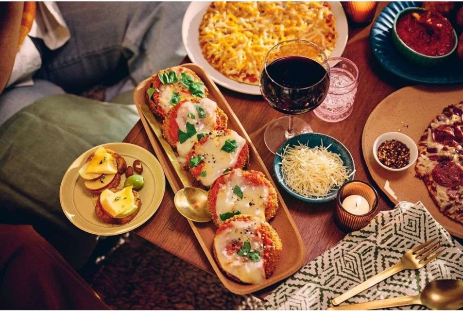 Overhead shot of a rustic wooden table featuring assorted pizzas, appetizers, red wine, and a lit candle, creating a cozy atmosphere.