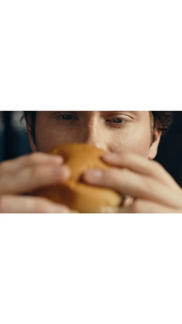 Close-up of an adult holding a burger. The person's eyes and hands are visible. The background is blurred and dark.