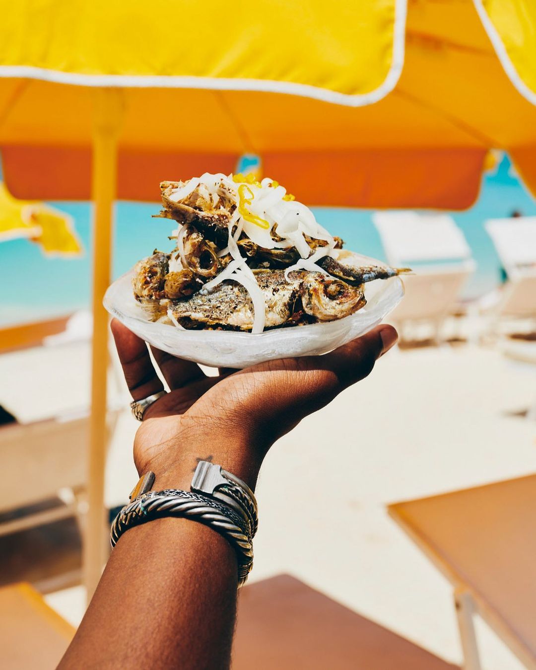 Close-up of an arm holding a plate of fried fish topped with shredded toppings against a bright beach backdrop with yellow umbrella.