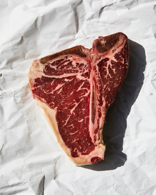 Overhead shot of a raw T-bone steak on white wrinkled parchment paper, showcasing marbling and bone structure, captured in bright light.