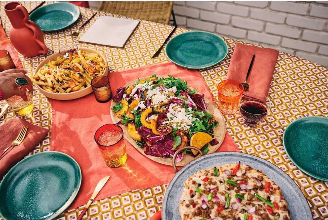 Overhead shot of a table set for a meal with pasta, salad, and pizza. Dinner plates, glasses, a water pitcher and flatware are set on table.