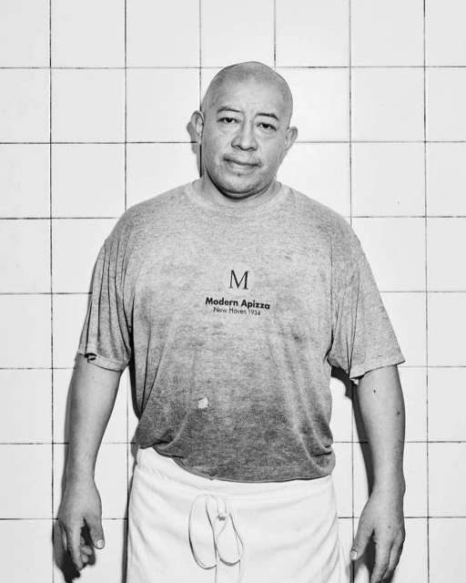 Black and white portrait of a man wearing a t-shirt and apron, standing indoors against a tiled wall, under bright studio lighting.