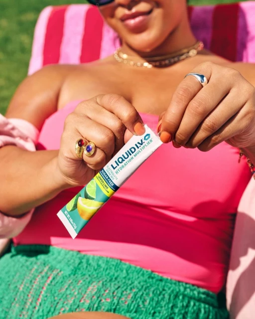 A woman sits in a lawn chair outside holding a Liquid I.V. hydration multiplier packet, wearing a pink top and sunglasses.